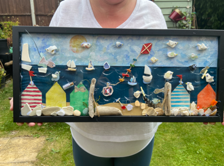 Beverley holding up 3D artwork of a beach scene with litter and other beach-combed objects incorporated.